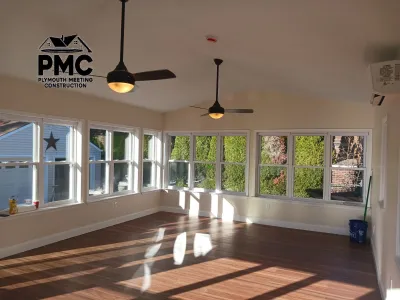 This bright, newly constructed sunroom features a vaulted ceiling, dual ceiling fans, extensive windows overlooking foliage, and warm wood-look flooring illuminated by natural light.