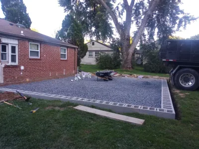A new gravel foundation pad, bordered by concrete blocks, is being constructed next to a red brick house in a suburban setting.