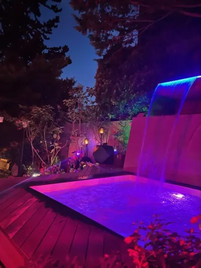 A luxurious backyard spa pool featuring a dramatic blue-lit waterfall and vibrant purple ambient lighting is surrounded by dark foliage and wooden decking at night.